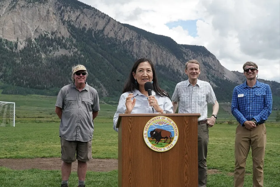 Sec. Deb Haaland speaking at dais outdoors with Colorado mountains in background and three officials standing behind her announcing protection of Thompson Divide.